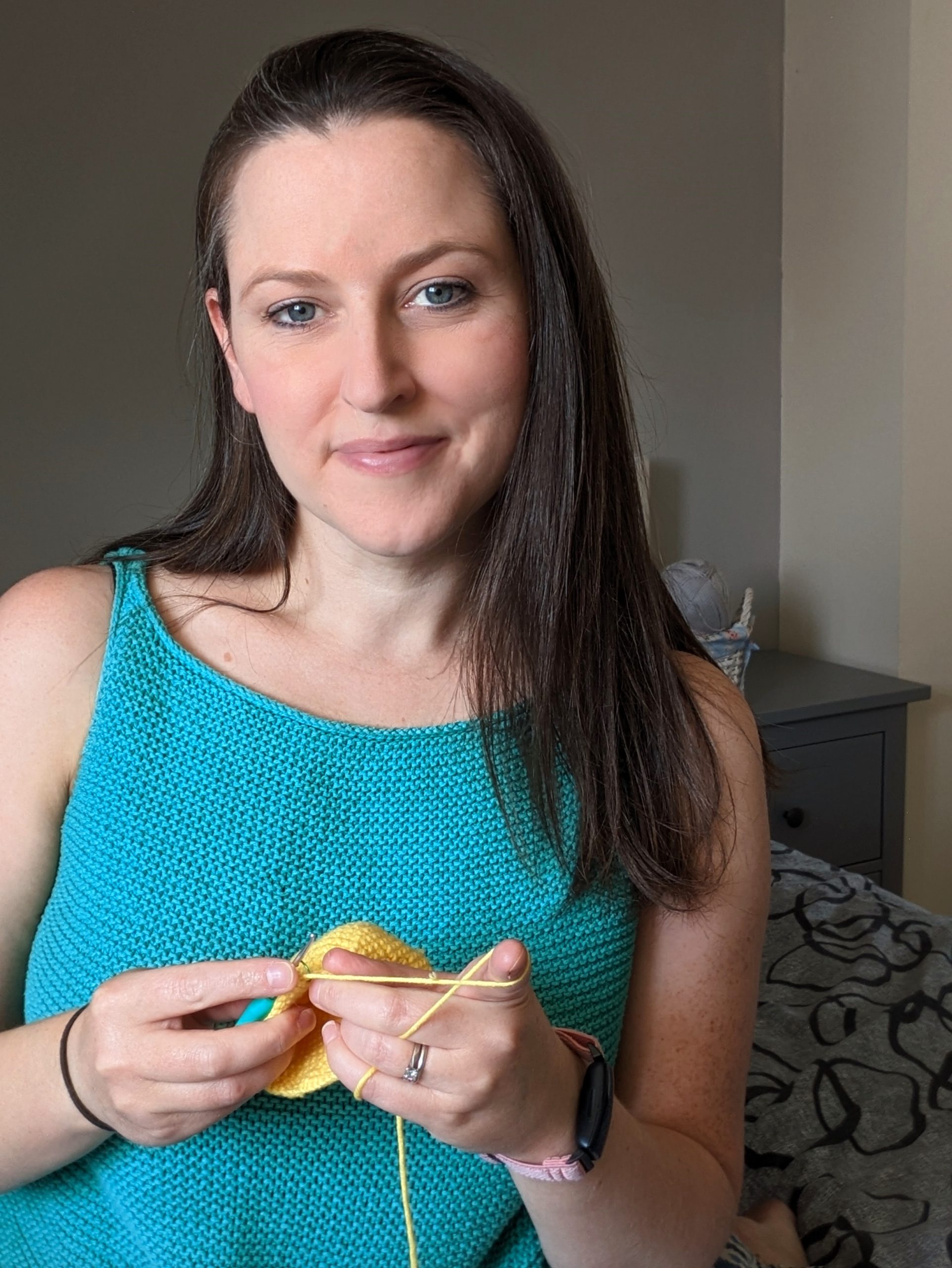 Megan is standing wearing a knitted top, holding yellow yarn and a crochet hook in her hands.