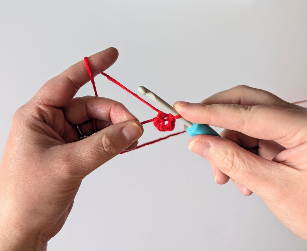 two hands holding yarn and a crochet hook, with the red yarn crocheted into a ring