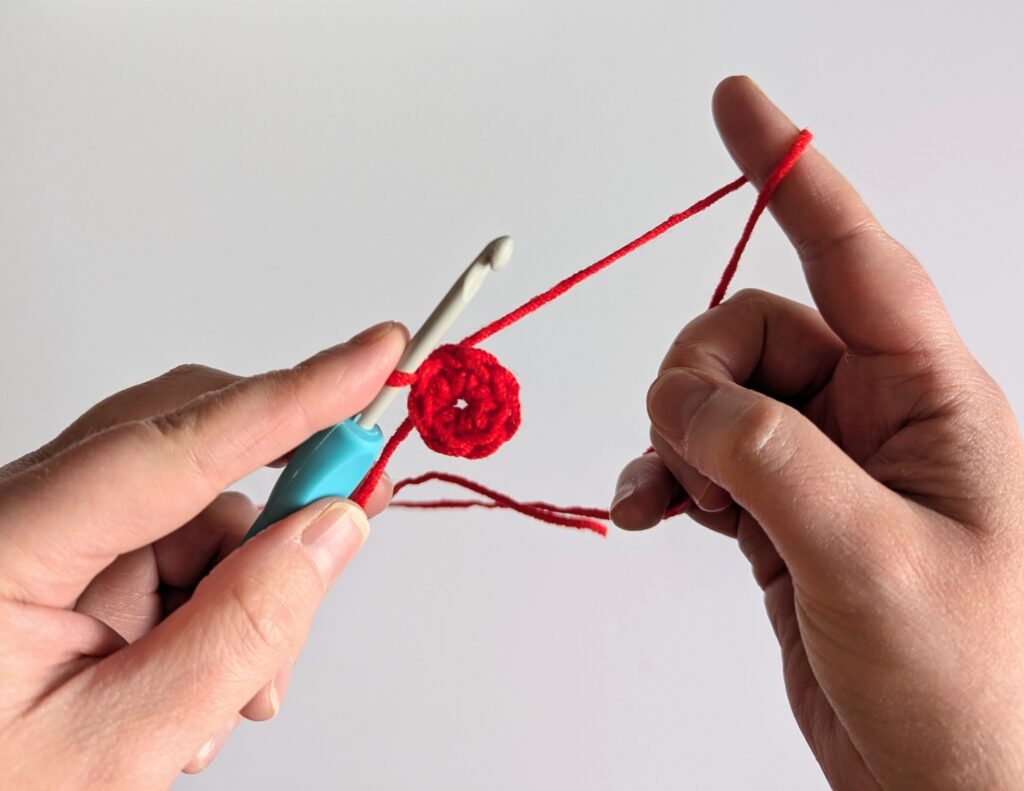 Two hands holding red yarn and a crochet hook, with a crocheted ring in the middle