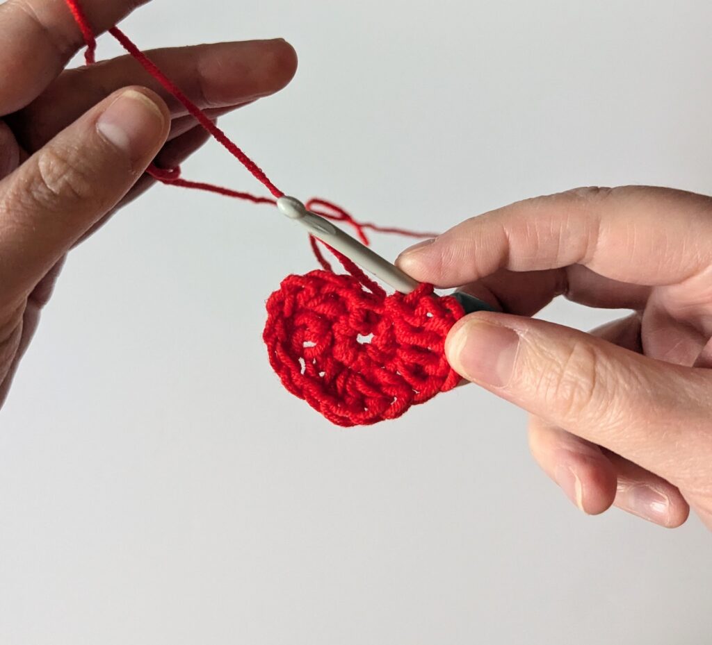 Two hands holding red yarn and a crochet hook, with a crocheted heart in the middle